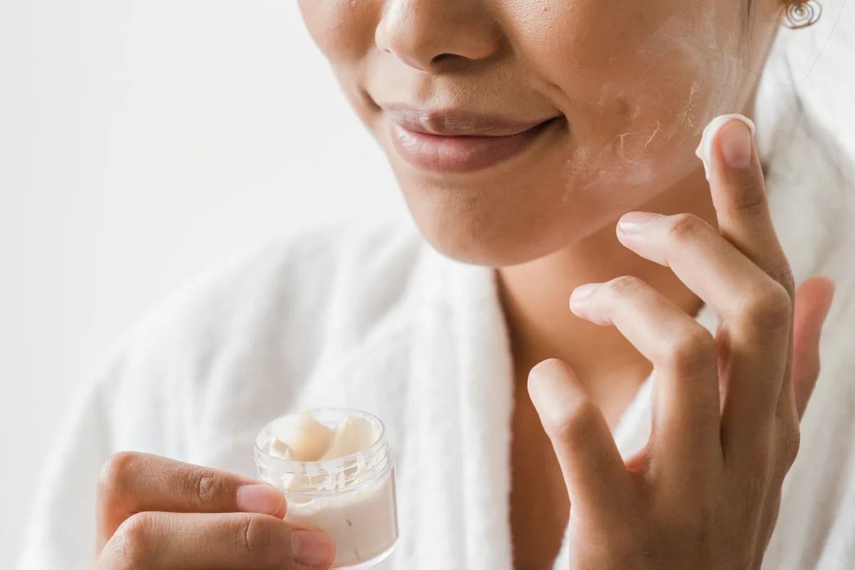 Imagen ilustrativa — Una mujer asiática adulta aplicando crema hidratante en el rostro, con bata blanca sobre un fondo claro